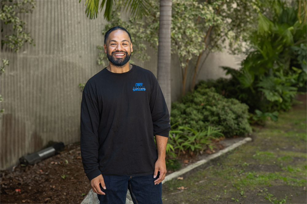 Brian Penesa stands outside in front of trees and plants along a concrete building wall