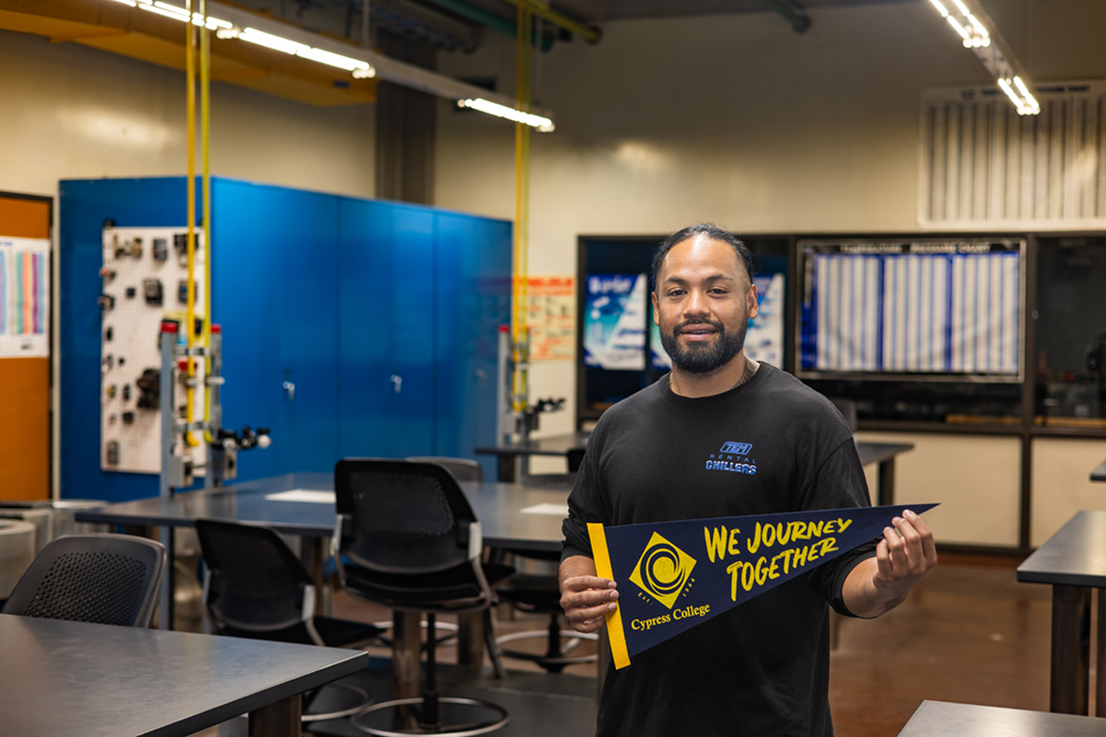 Brian Penesa holds a Cypress College pennant inside an Air Conditioning and Refrigeration classroom.